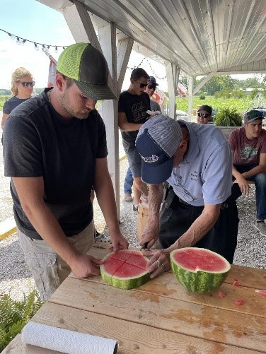 Two people cutting a watermelon open on a wooden table outdoors